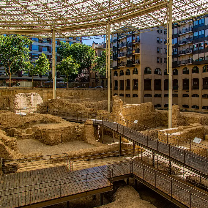 Ruinas arqueológicas bajo un techo moderno, rodeadas de edificios y árboles.