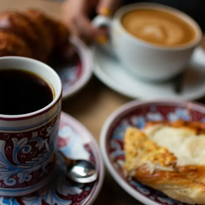 Tazas de café y platillos con croissants y pasteles sobre una mesa.