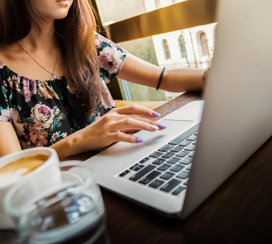 Mujer usando una laptop en una cafetería con una taza de café y ventana de fondo.