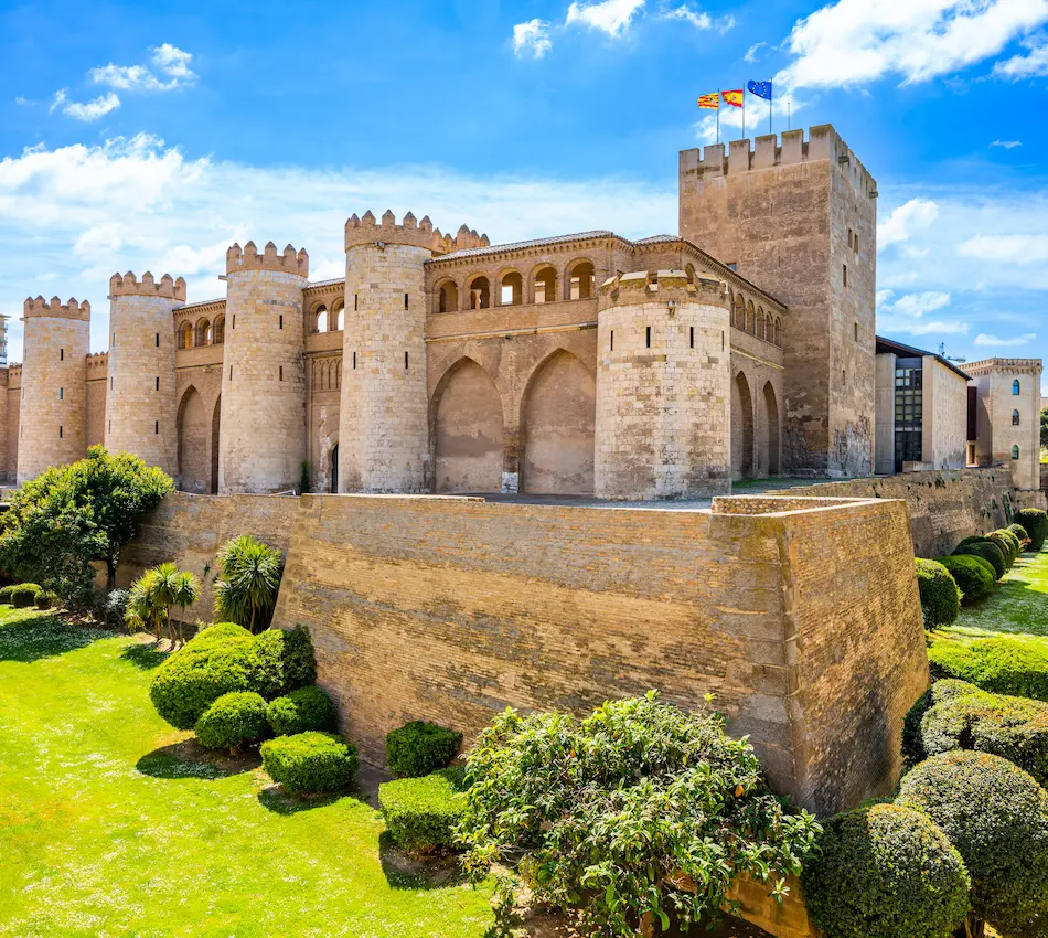 Antiguo castillo con torres y murallas, rodeado de jardines y bajo un cielo azul.