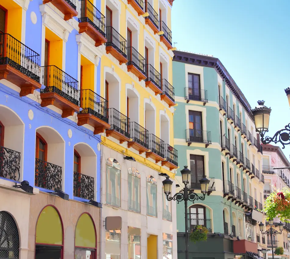 Edificios coloridos con balcones y arcos en una calle, con cielo azul y faroles antiguos.