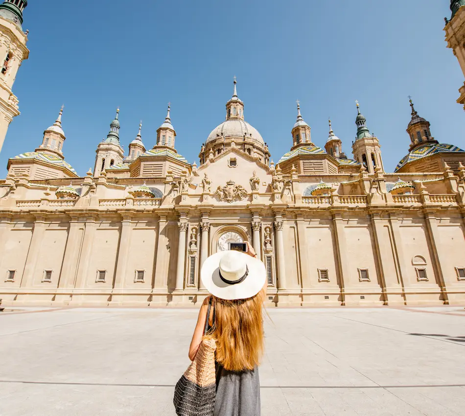 Mujer con sombrero, de espaldas, fotografía una catedral con múltiples cúpulas bajo un cielo despejado.