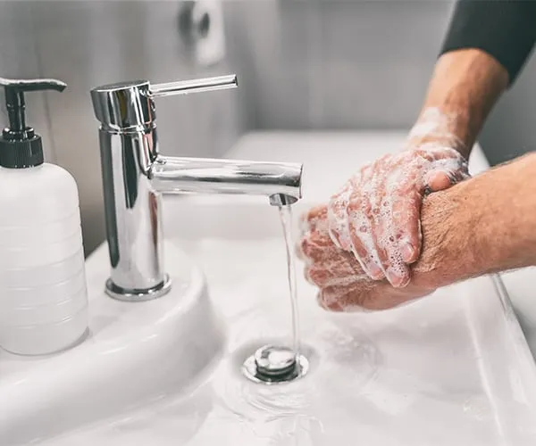 Manos lavándose con agua y jabón en un lavabo, junto a un dispensador de jabón líquido.