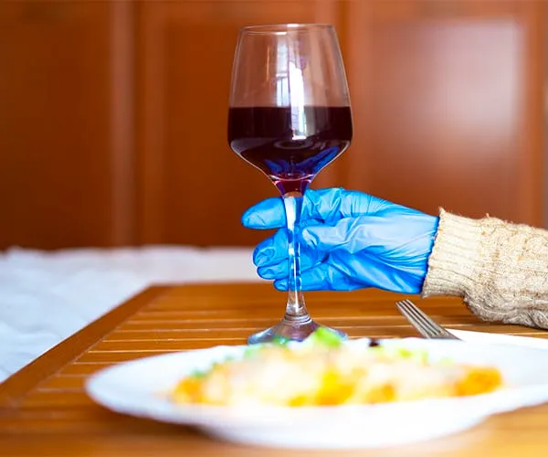 Mano con guante azul sosteniendo una copa de vino tinto junto a un plato de comida sobre una mesa.