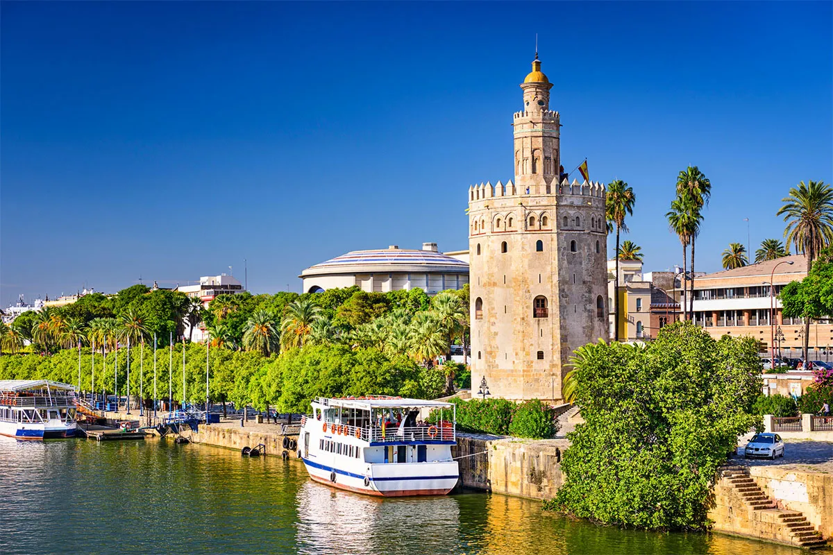 Torre junto al río con barcos y vegetación. Cielo azul despejado.