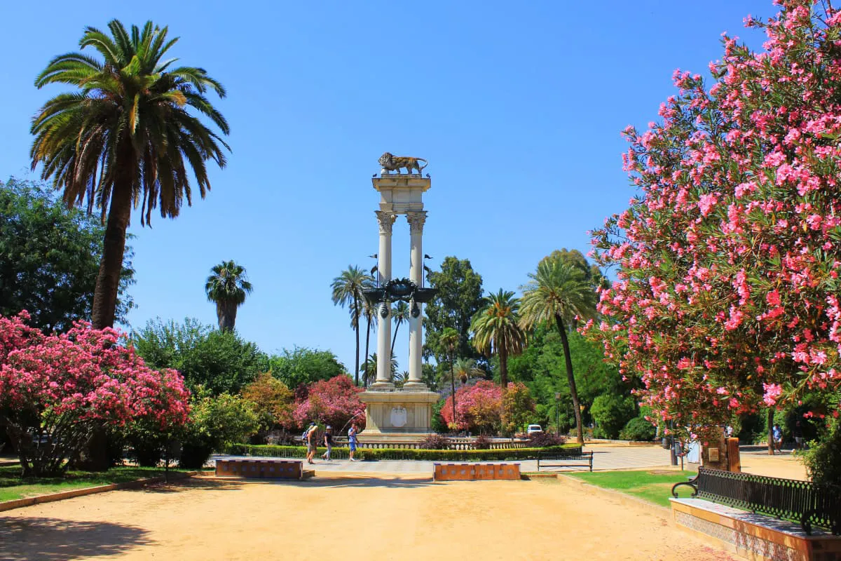 Parque con palmeras, flores rosas y monumento con columnas coronadas por una escultura de león.