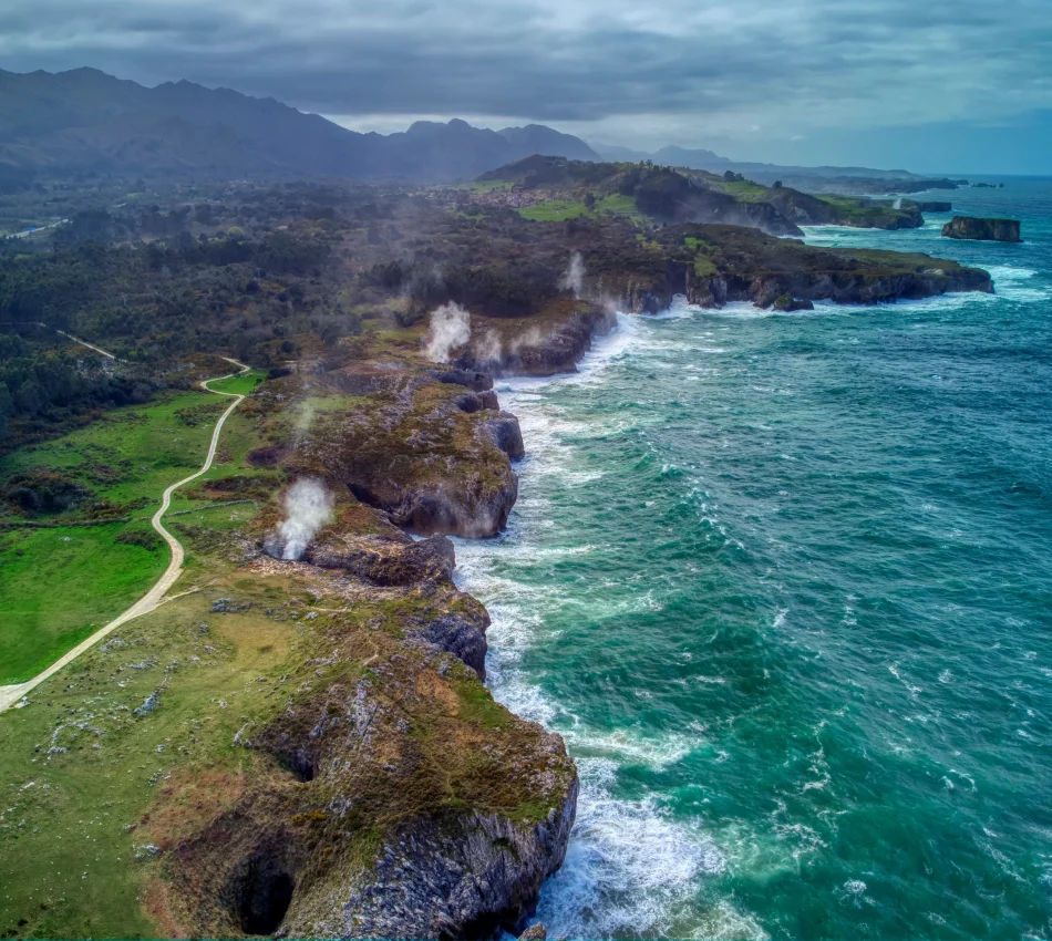 Acantilados junto al mar con olas, sendero y columnas de humo ascendentes.