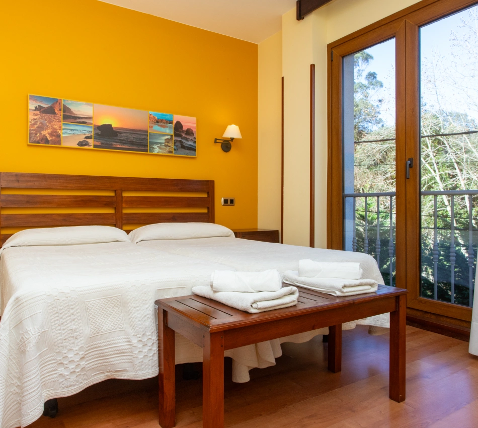 Habitaci&oacute;n con dos camas, pared amarilla, ventanal y cuadros de paisajes.