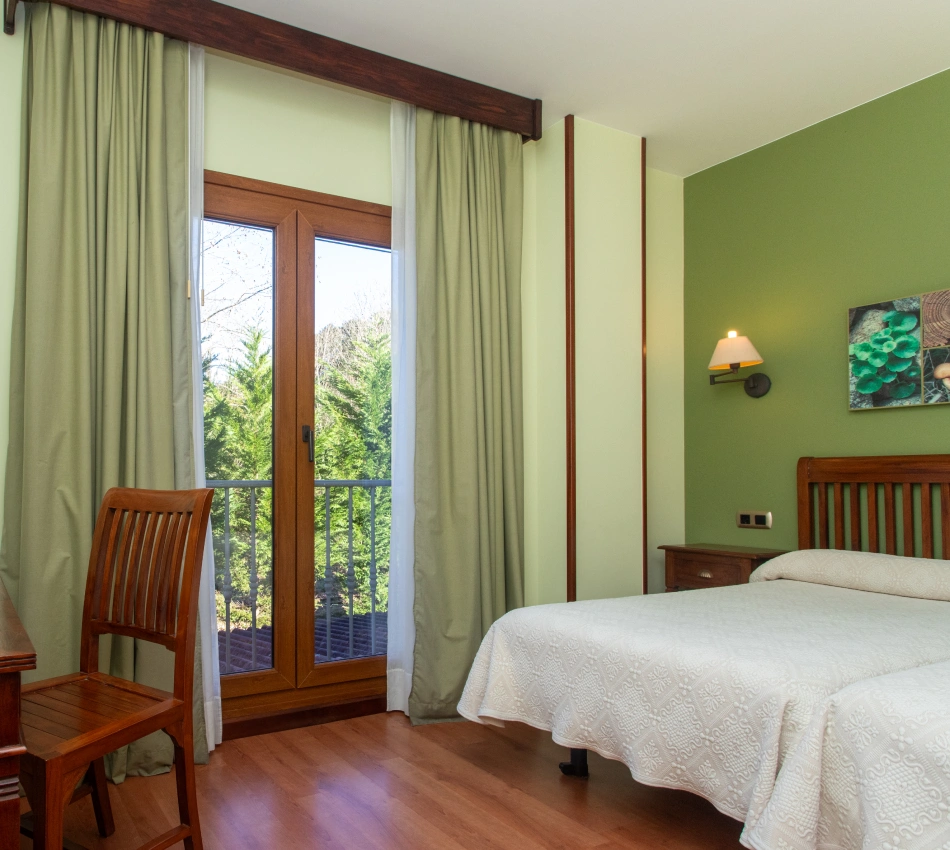 Habitaci&oacute;n con cama doble, silla de madera, ventana grande y paredes verde claro.
