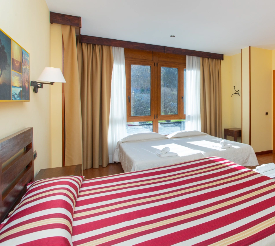 Habitaci&oacute;n con dos camas, cortinas beige, ventana amplia y cuadros en la pared.