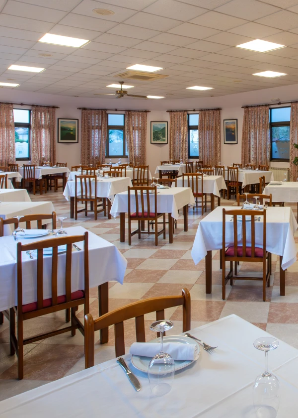 Comedor vac&iacute;o con mesas cuadradas, sillas de madera y manteles blancos. Cortinas estampadas.
