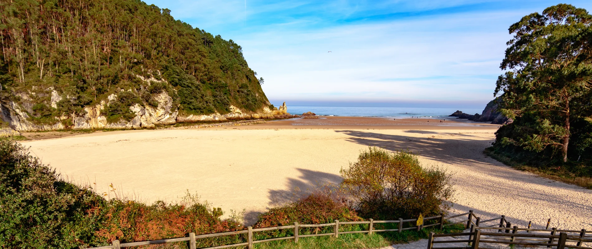 Bah&iacute;a tranquila con playa de arena, rodeada de acantilados verdes y cielo azul.