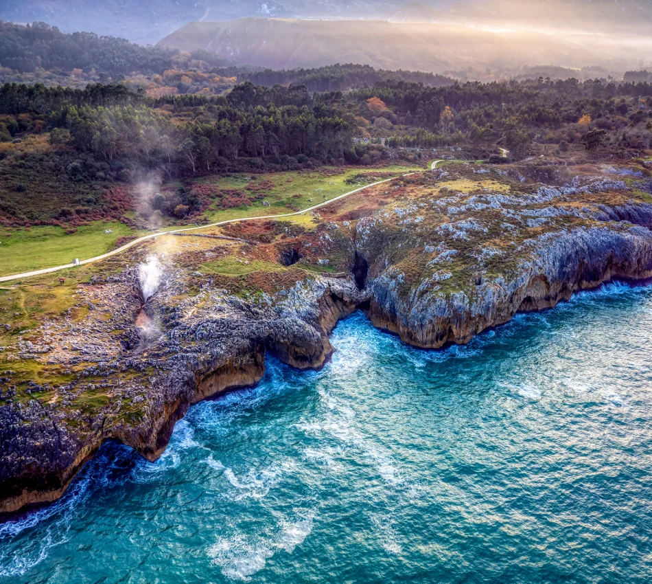 Acantilados junto al mar con vegetaci&oacute;n, vapor y un camino. Monta&ntilde;as al fondo, paisaje costero.