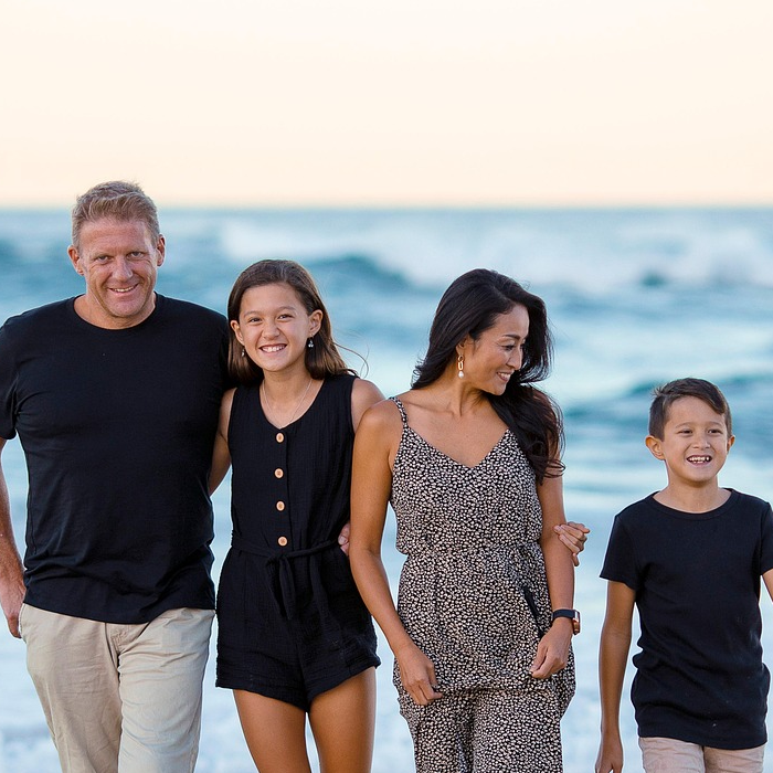 Familia caminando por la playa, sonrientes y abrazados, con el mar de fondo al atardecer.