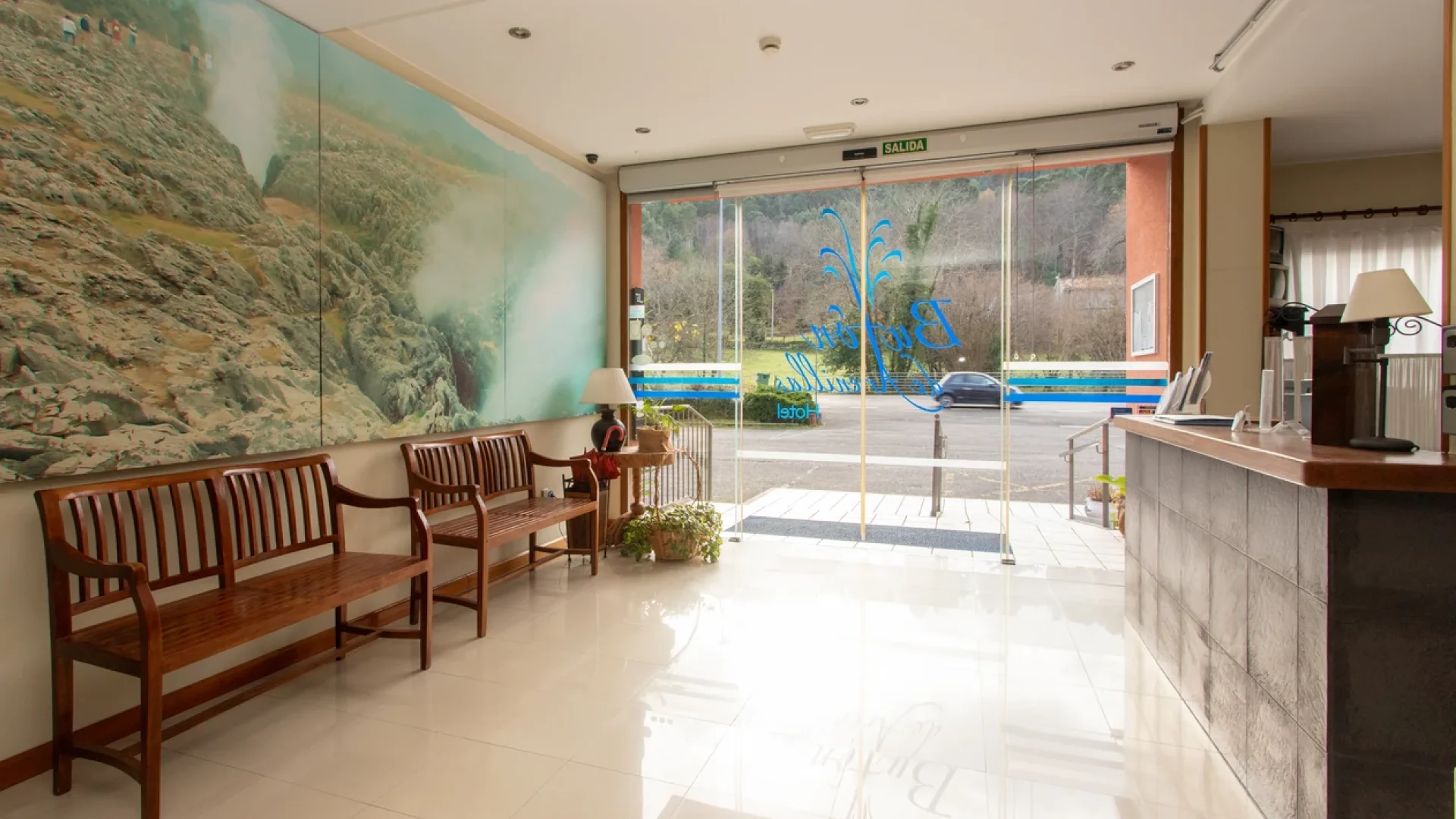 Vest&iacute;bulo de hotel con bancos de madera, gran mural y puertas de vidrio que dan a la calle.