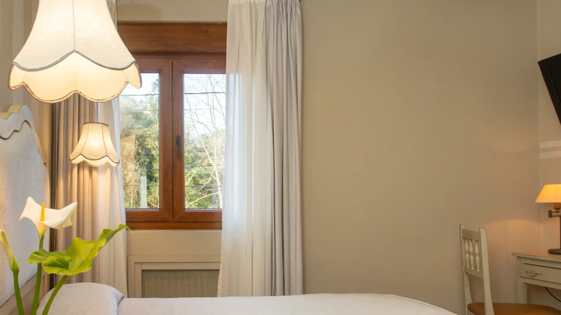 Habitaci&oacute;n con ventana, cortinas blancas, l&aacute;mparas y flores junto a la cama.