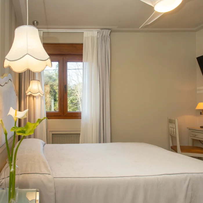 Habitaci&oacute;n luminosa con cama blanca, ventana grande, l&aacute;mpara de mesa y flor sobre la mesa.