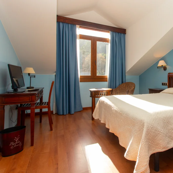 Habitaci&oacute;n luminosa con cama, escritorio, televisor y ventana con cortinas azules.
