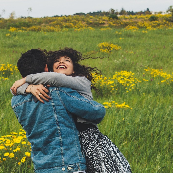 Pareja abraz&aacute;ndose y girando en un campo lleno de flores amarillas y hierba verde.