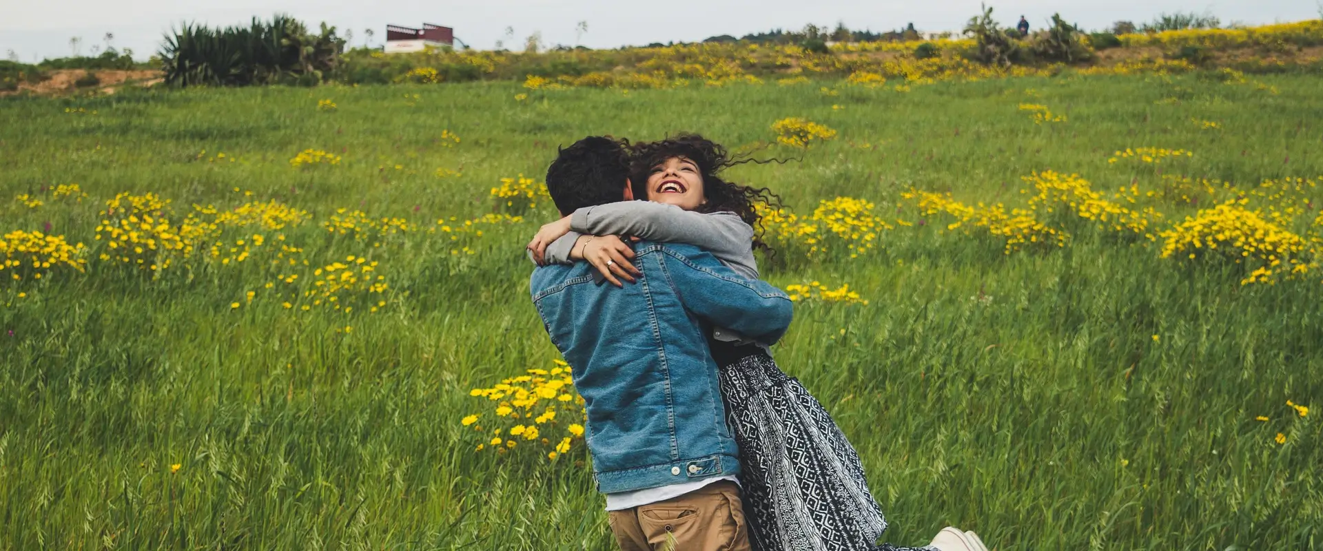 Pareja abraz&aacute;ndose alegremente en un campo verde con flores amarillas.