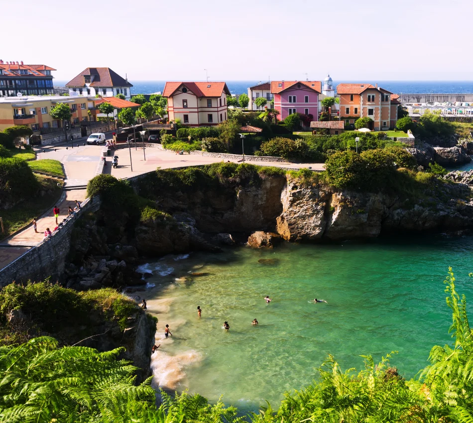 Casas coloridas y acantilado sobre playa turquesa con ba&ntilde;istas nadando, vegetaci&oacute;n en primer plano.