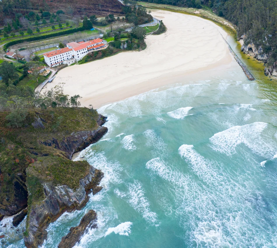 Vista a&eacute;rea de una playa con un edificio blanco y tejado rojo al fondo, rodeada de vegetaci&oacute;n.