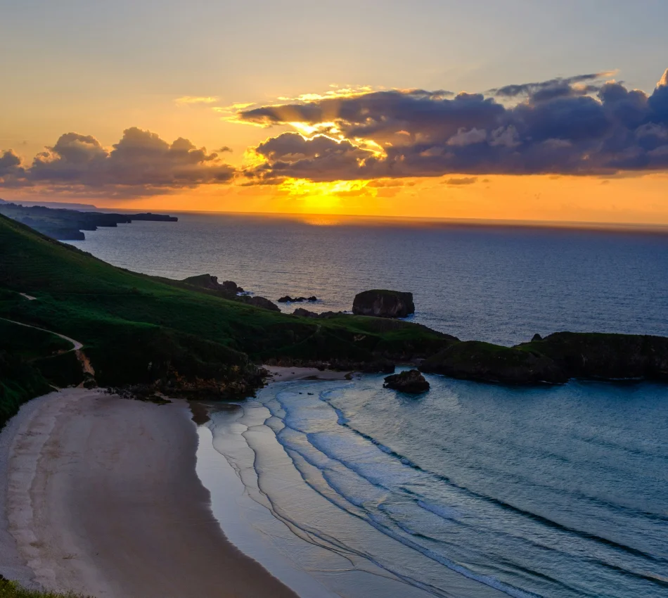 Atardecer en una playa con olas suaves, acantilados verdes y un cielo nublado.