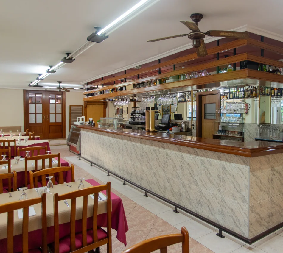 Interior de un restaurante con sillas de madera y un bar con botellas y copas colgantes.