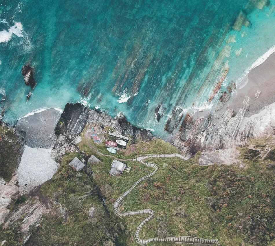 Vista a&eacute;rea de costa rocosa con mar turquesa, camino sinuoso y peque&ntilde;as estructuras en la hierba.