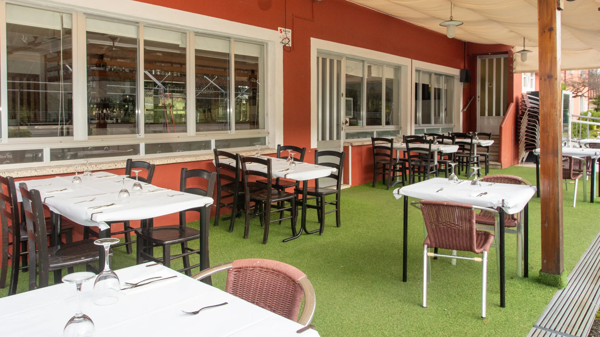 Terraza de restaurante con mesas y sillas sobre c&eacute;sped artificial, junto a un edificio rojo.