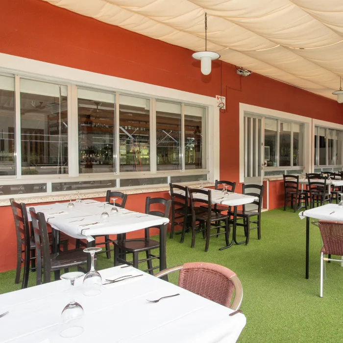 Terraza cubierta con mesas y sillas, c&eacute;sped artificial y fondo rojo con ventanas.