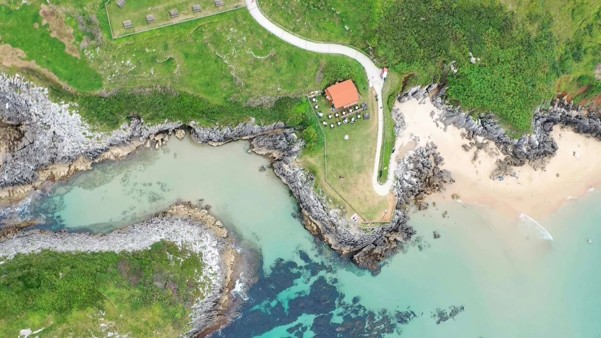 Vista a&eacute;rea de un acantilado verde con playa, mar azul y peque&ntilde;a construcci&oacute;n con techo rojo.