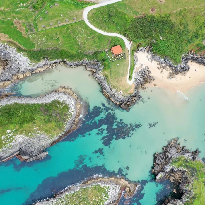 Vista a&eacute;rea de paisaje costero con playa, rocas y sendero rodeado de vegetaci&oacute;n verde.