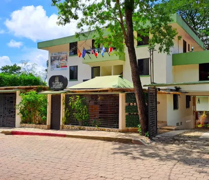 Edificio de tres plantas con banderas y vegetaci&oacute;n, visto desde la calle en un d&iacute;a soleado.