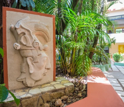 Relieve maya en un jard&iacute;n con plantas y arquitectura moderna al fondo.