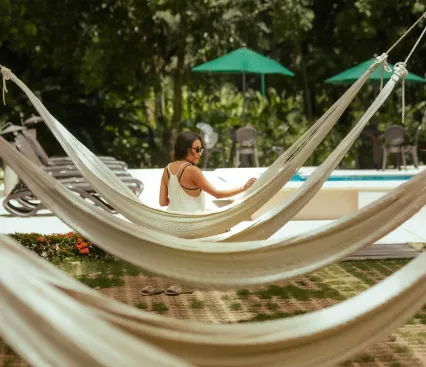 Mujer en hamaca al aire libre junto a una piscina rodeada de &aacute;rboles y sombrillas verdes.