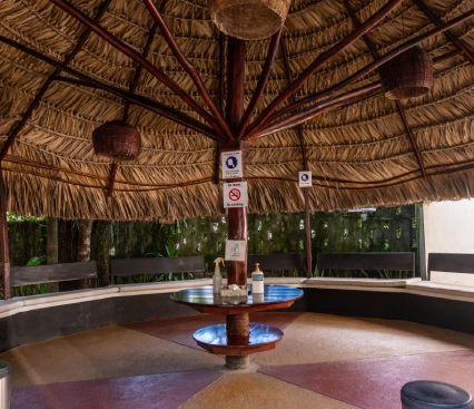 &Aacute;rea de descanso circular con techo de paja y bancos, mesas al centro, decorado r&uacute;stico.