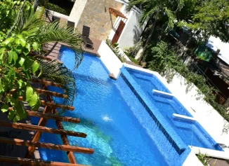 Piscina azul rodeada de vegetaci&oacute;n con p&eacute;rgola de madera y tumbonas al fondo.