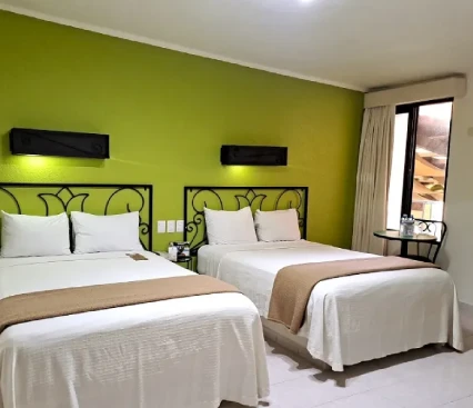 Habitaci&oacute;n de hotel moderna con dos camas, pared verde y ventana con cortinas claras.