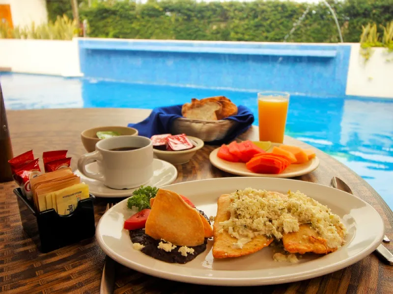 Desayuno junto a la piscina: caf&eacute;, pan, fruta, jugo y plato principal con queso y frijoles.