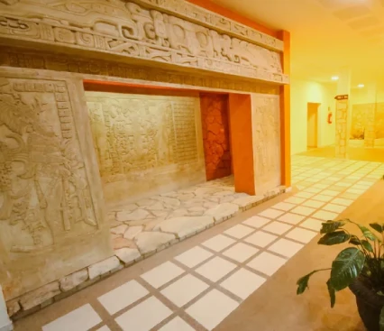 Pasillo con relieves arqueol&oacute;gicos en la pared y una planta decorativa al frente.