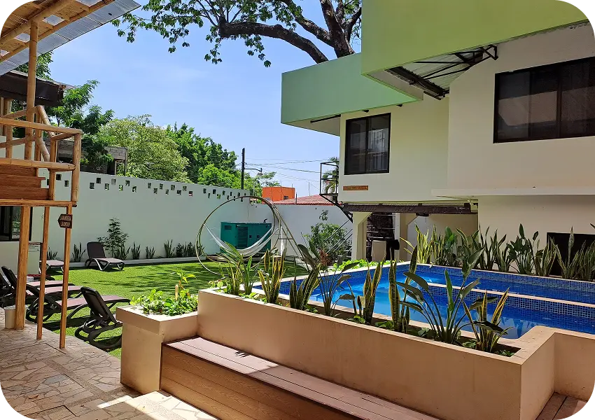 &Aacute;rea exterior con piscina, tumbonas, columpio y vegetaci&oacute;n, junto a un edificio moderno.