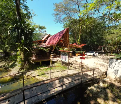 Puente y caba&ntilde;as con techo rojo rodeados de &aacute;rboles y vegetaci&oacute;n en un entorno soleado.
