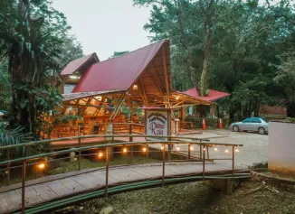Caba&ntilde;a con techo rojo, puente y &aacute;rboles alrededor, ambiente natural y estacionamiento con un auto.