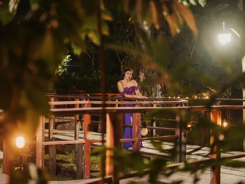 Mujer con vestido lila en un puente de madera, entre hojas y luz de farola nocturna.