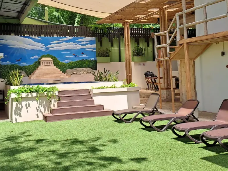 Terraza con c&eacute;sped artificial, tumbonas y mural de una pir&aacute;mide maya rodeada de plantas.