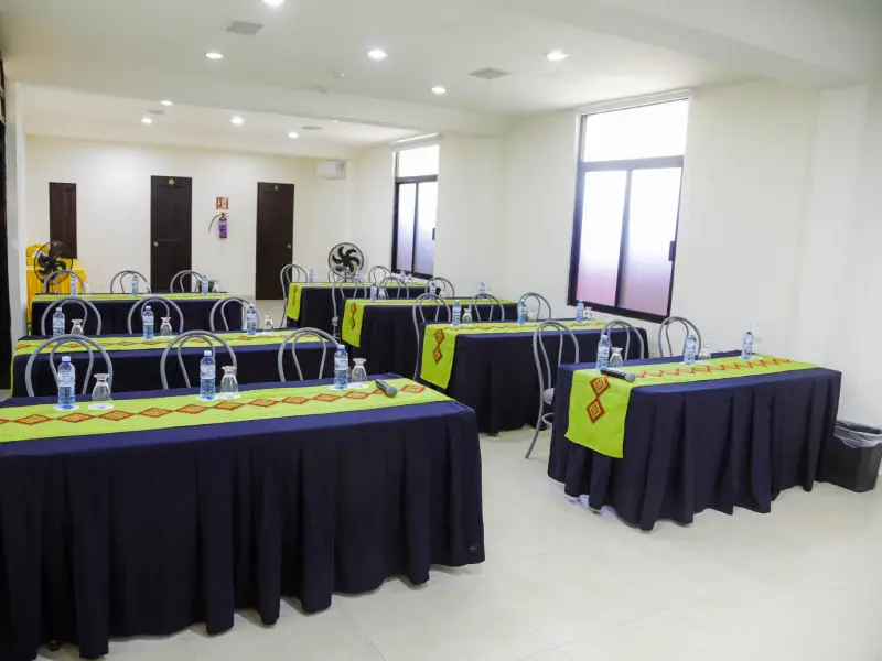 Sala de reuniones con mesas y sillas alineadas, decoradas con manteles verdes y botellas de agua.