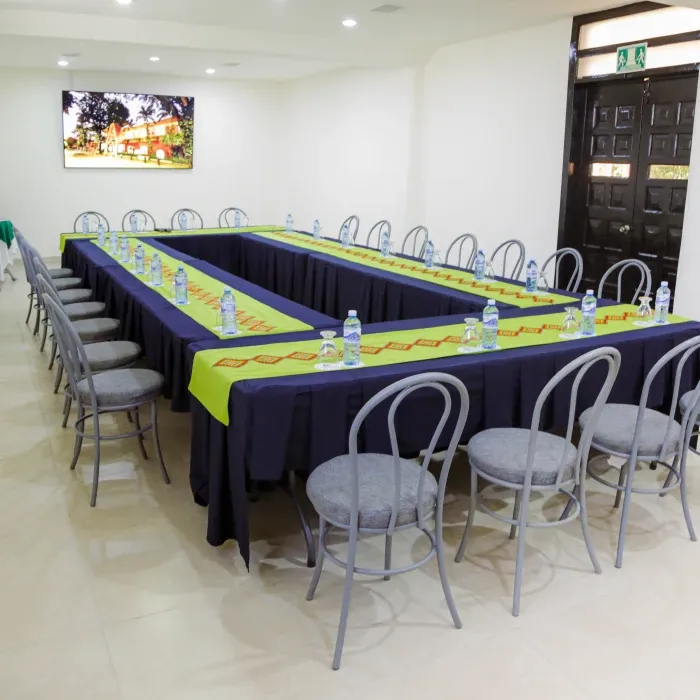 Sala de reuniones con mesas en forma de U, manteles verdes y botellas de agua sobre ellas.