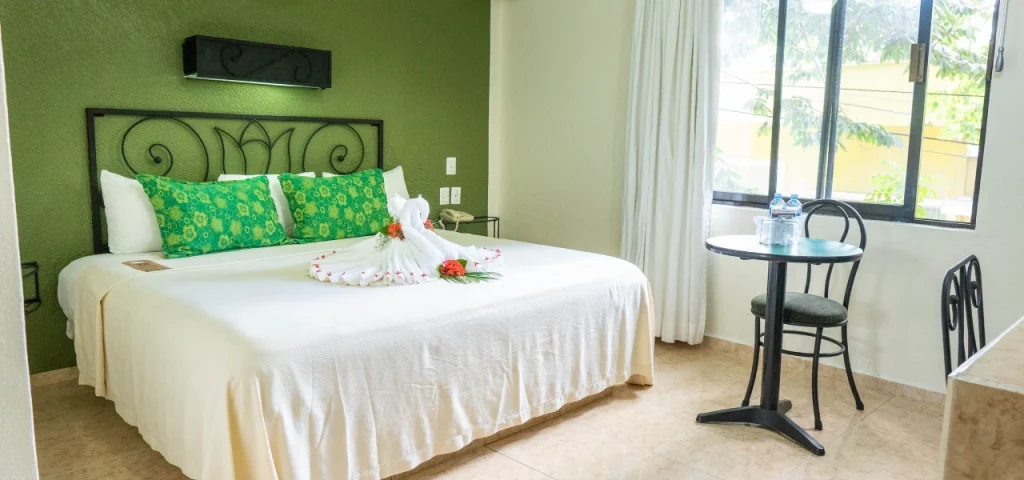 Habitaci&oacute;n de hotel con cama decorada, mesa peque&ntilde;a y ventana con cortinas blancas.