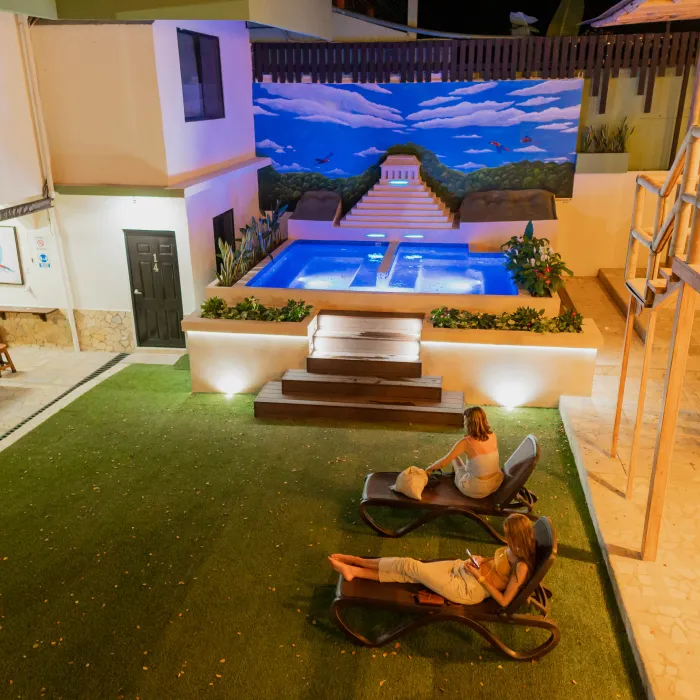 Dos personas descansando en tumbonas frente a una piscina iluminada, con un mural al fondo.